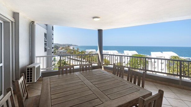 Ein Balkon mit Holztisch und -stühlen mit Blick auf die Küste, Gebäude und das Meer.