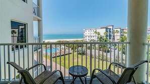 A balcony with a view of the beach and pool, featuring outdoor furniture and a clear sky.