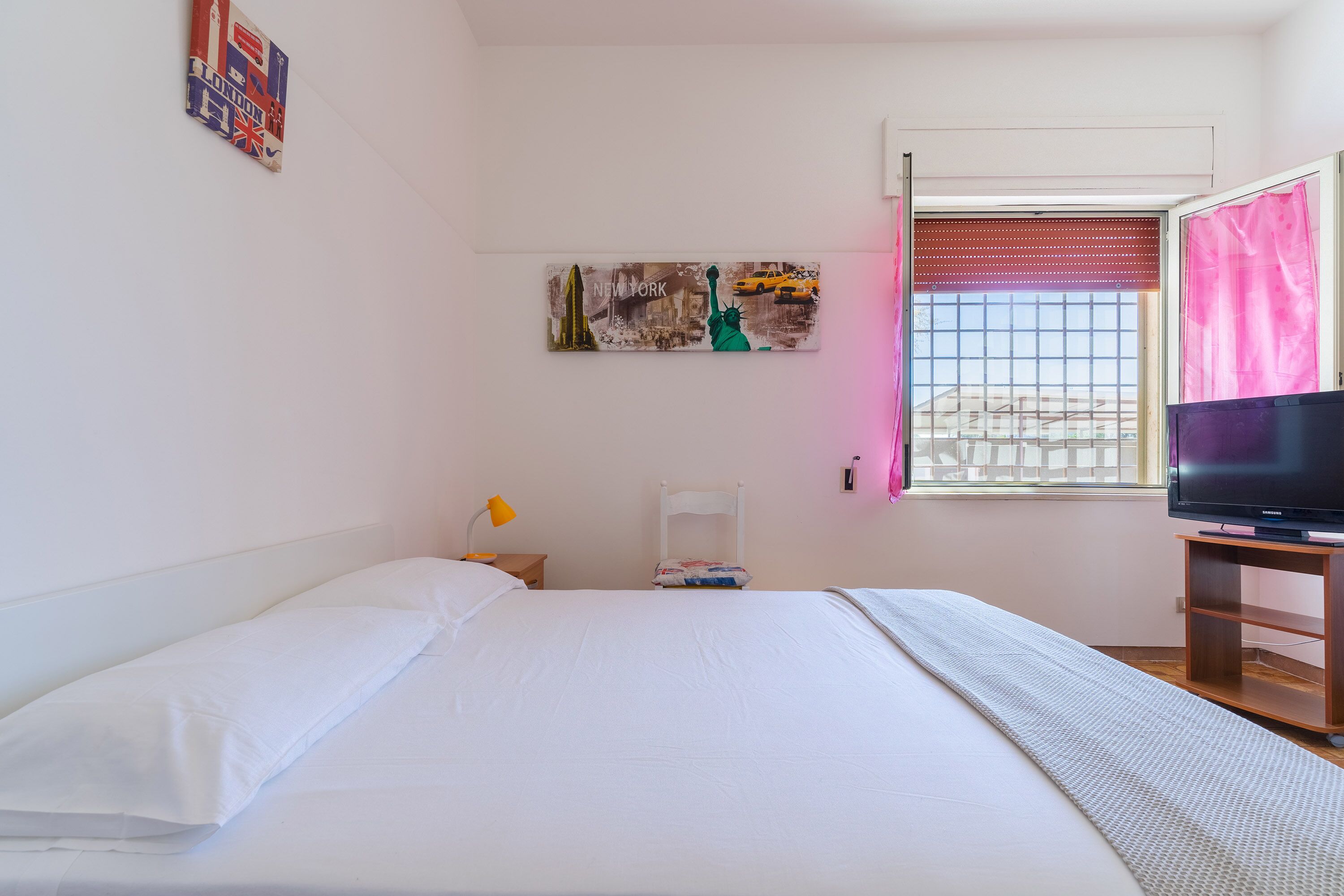 Un dormitorio con cama, televisor sobre un mueble, ventana con persianas y una obra de arte en la pared.