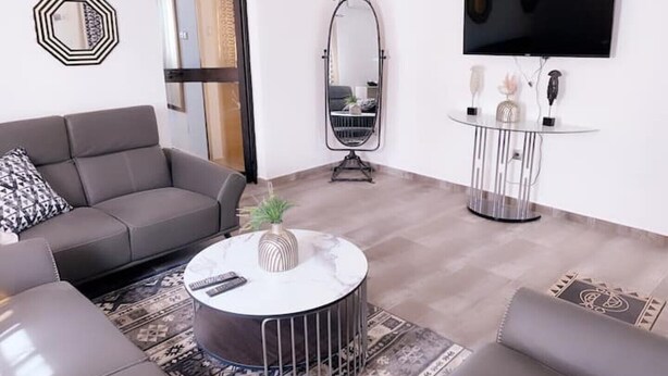 Una sala de estar moderna con un sofá gris, una mesa redonda con un florero, un televisor de pantalla plana y un espejo decorativo.
