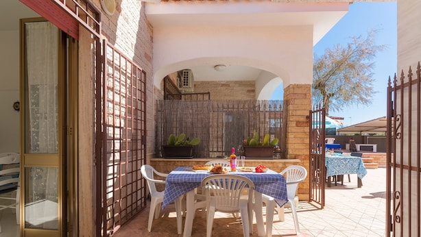 An outdoor patio with a table set for dining, surrounded by white chairs, and a brick wall in the background.
