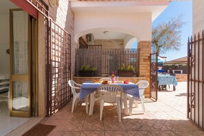 Um pátio externo com uma mesa posta para refeições, rodeado por cadeiras brancas, e um muro de tijolos ao fundo.