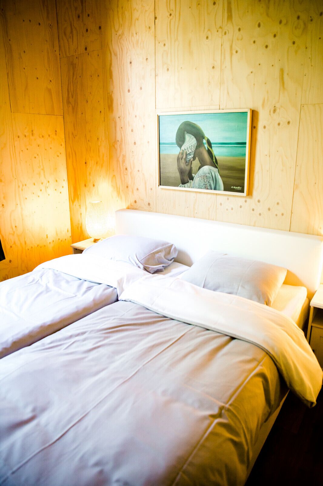 Ett hotellrum med en väggpanel av trä, en säng med vita sängkläder, ett nattduksbord med en lampa och en inramad tavla på väggen.