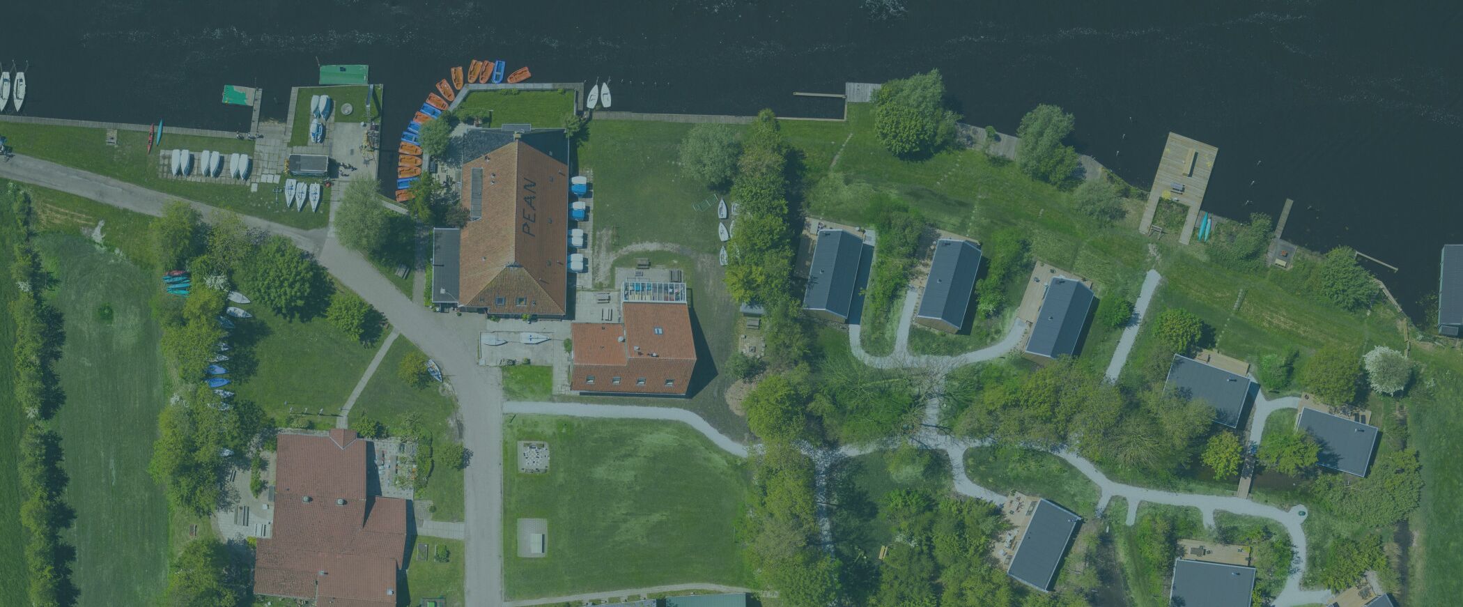 Une vue aérienne d’une zone côtière, avec un grand bâtiment portant l’inscription « PEAN » et plusieurs structures plus petites, le tout entouré de verdure et bordé par une étendue d’eau.