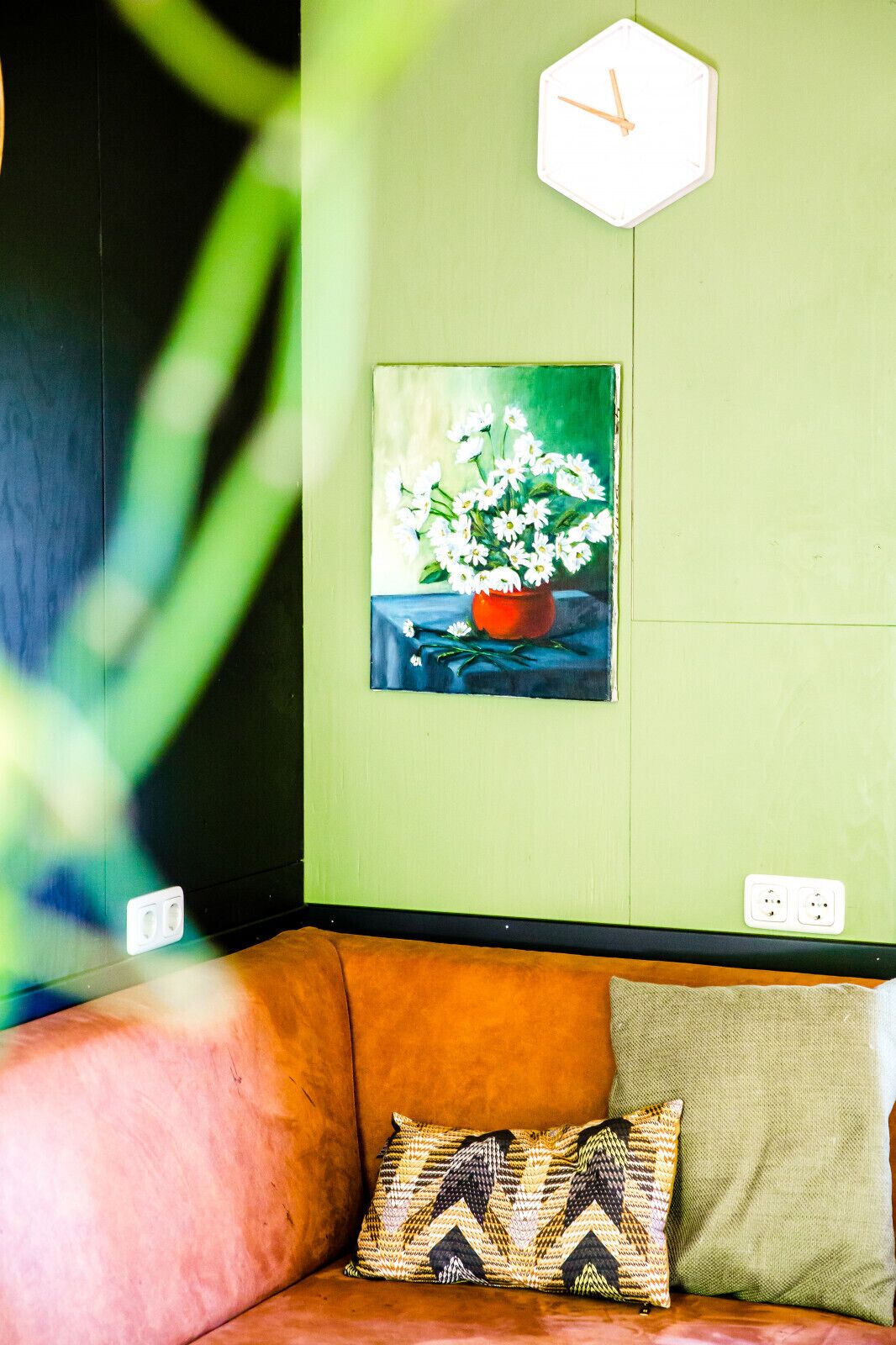 Et værelse med grøn væg, et ur, et blomsterbillede, en brun sofa og en pude med mønster.