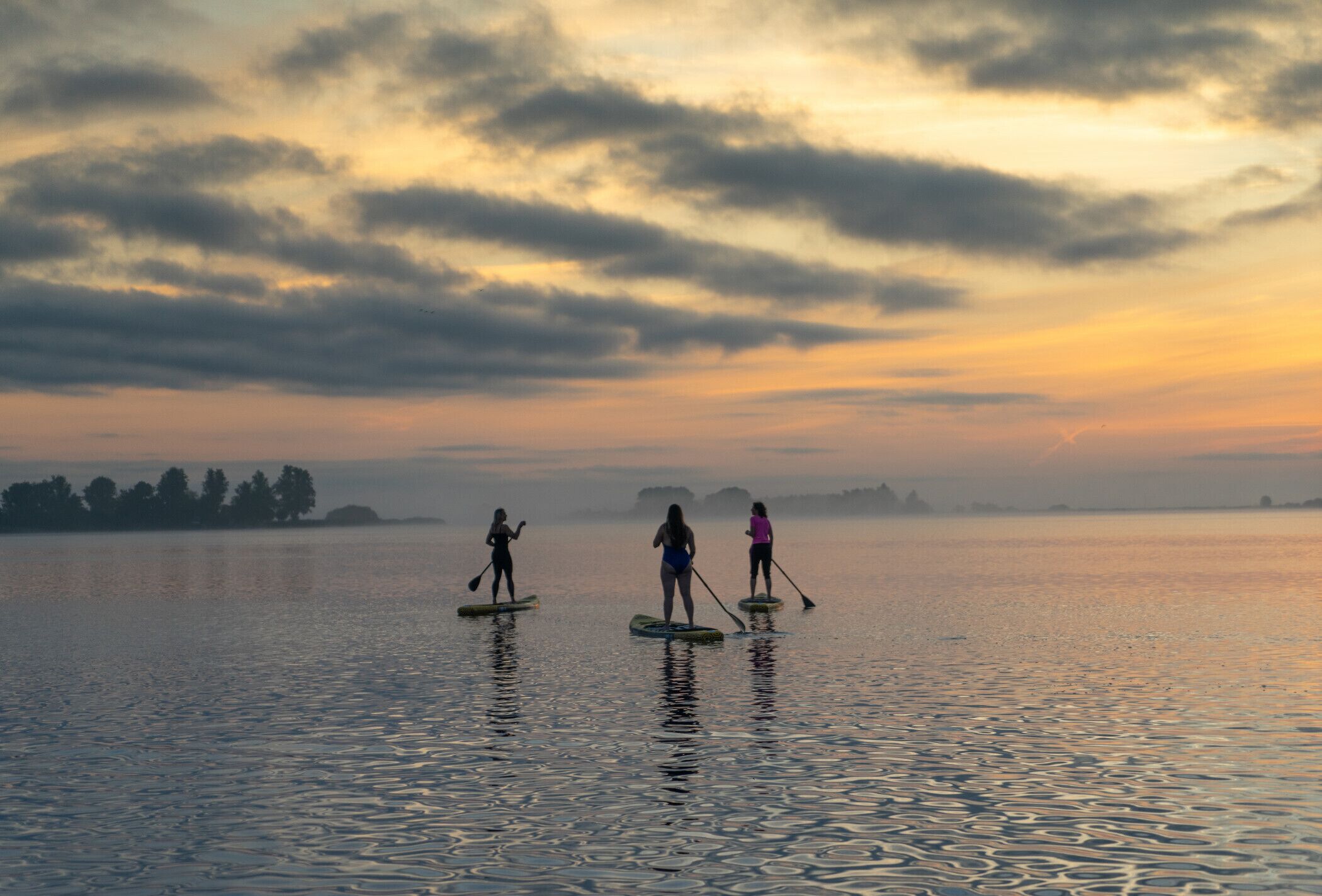 Three people paddleboarding on a calm lake during sunset.