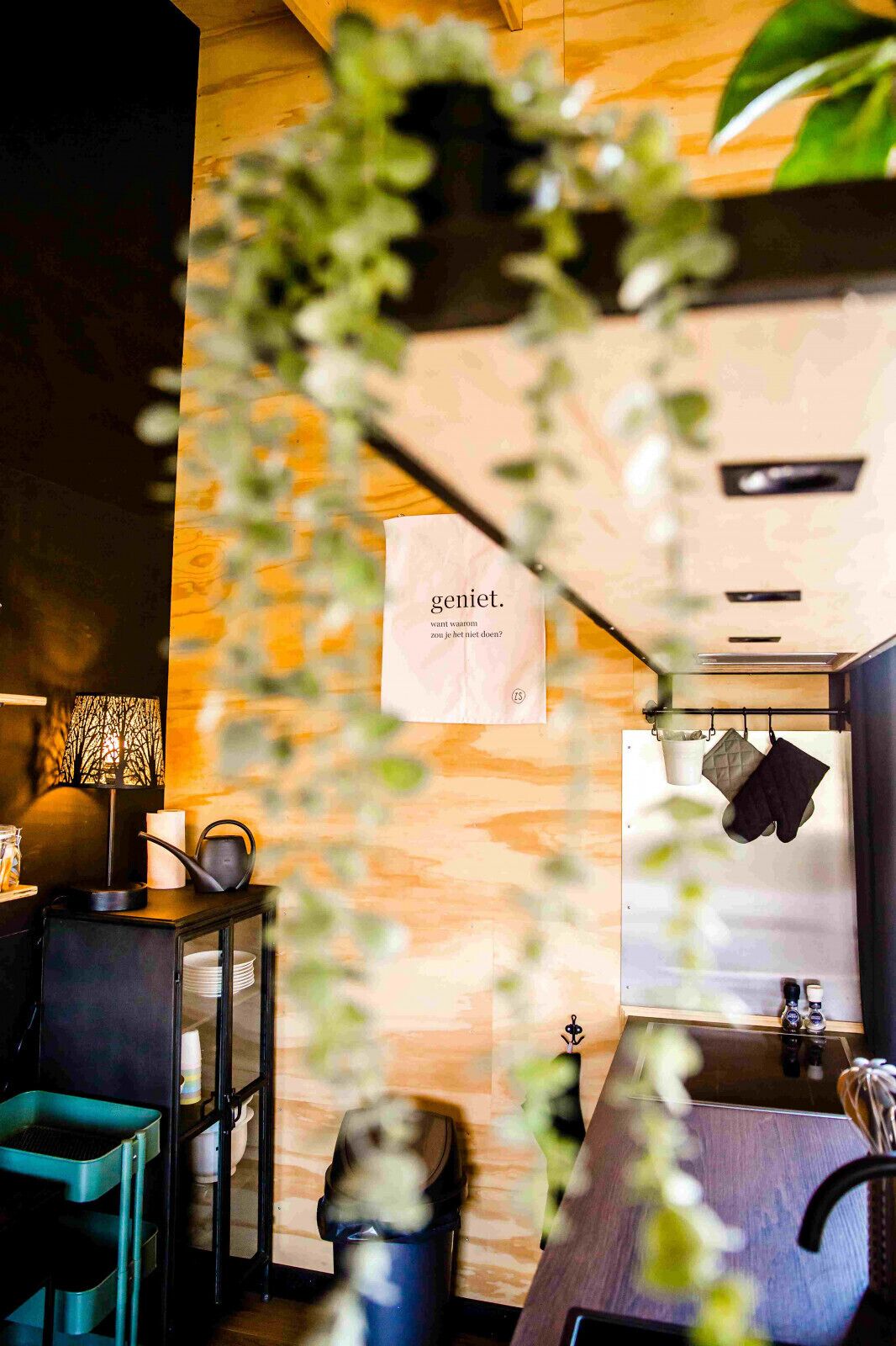 A modern kitchen with wooden cabinetry, a hanging pot rack, and a visible trash bin.