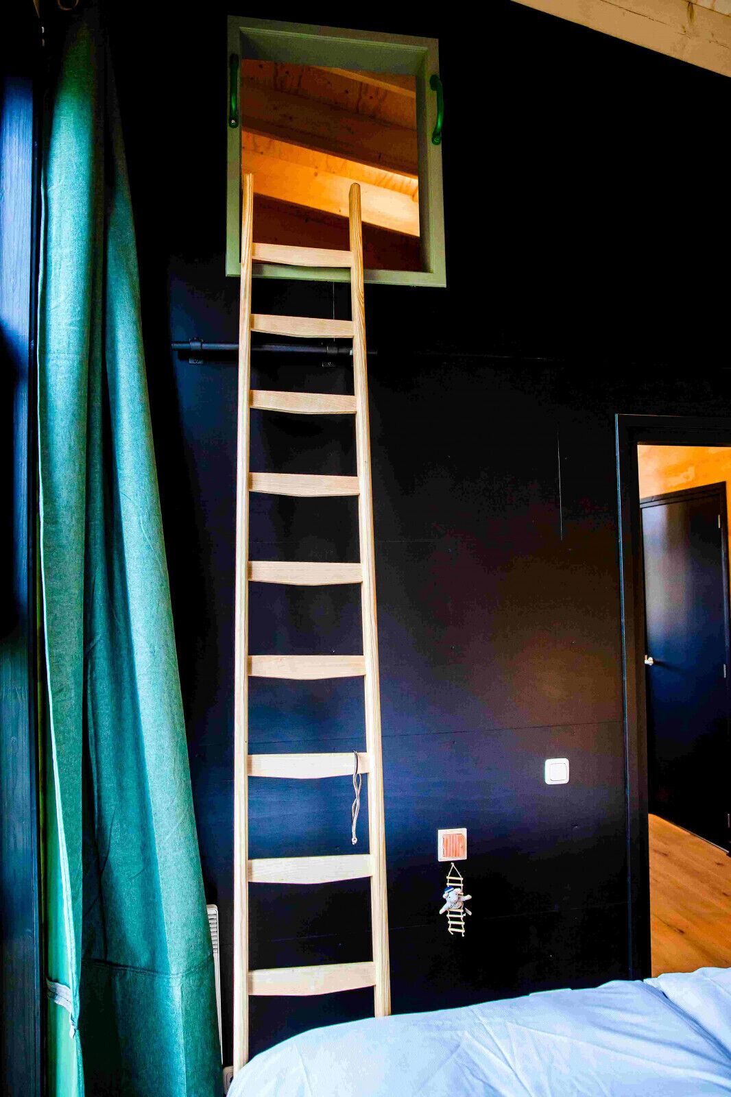 A room with a wooden ladder against a dark wall, a bed with white bedding, and a green curtain.