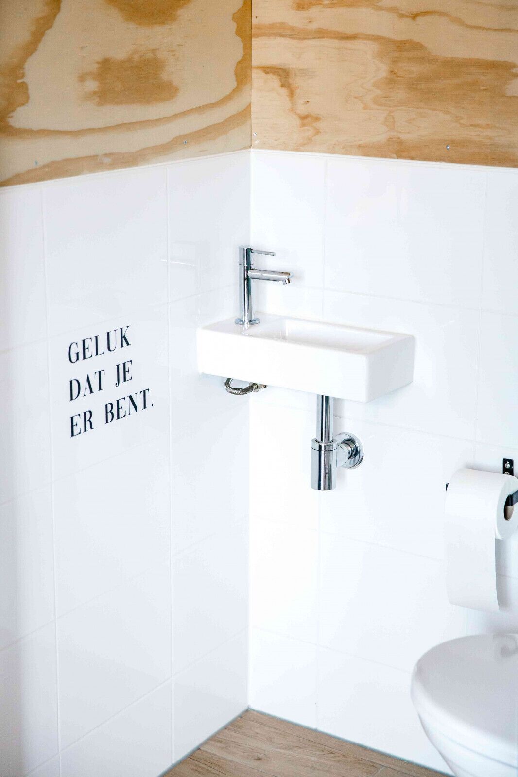 Une salle de bain avec un lavabo blanc, des toilettes et un mur dont une partie est recouverte de carreaux.