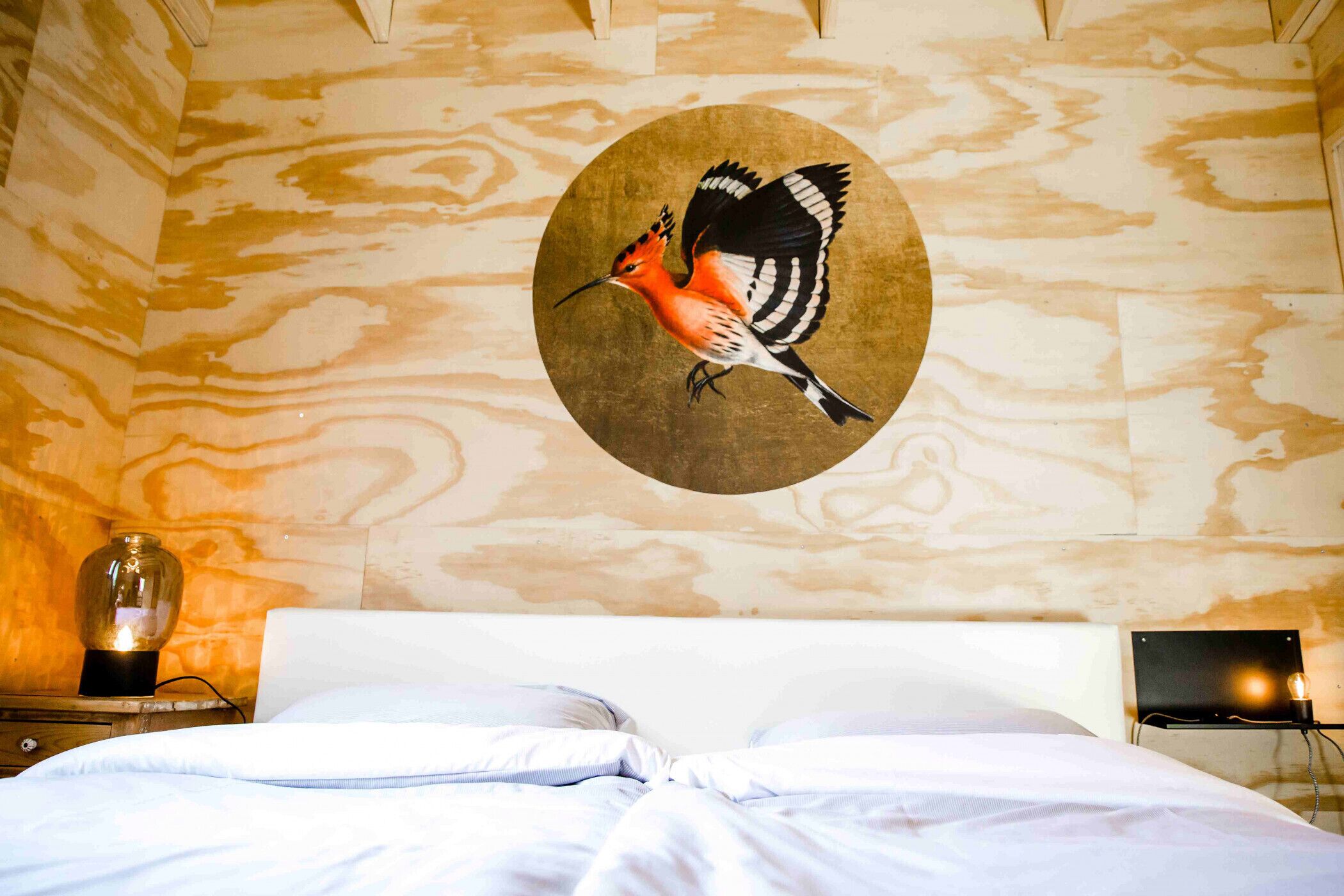A room with a bed, a wall-mounted circular artwork of a bird, a nightstand with a lamp, and a wall-mounted light fixture.