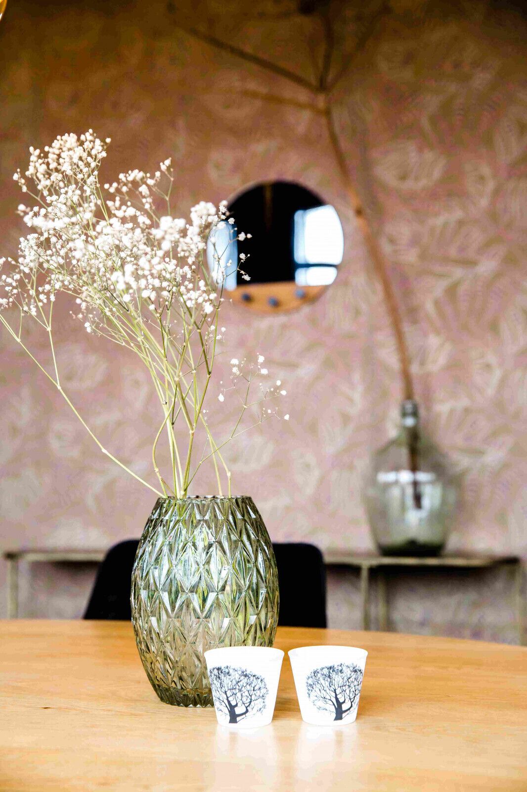 A table with a textured vase containing white flowers, two cups with tree designs, and a mirror in the background.