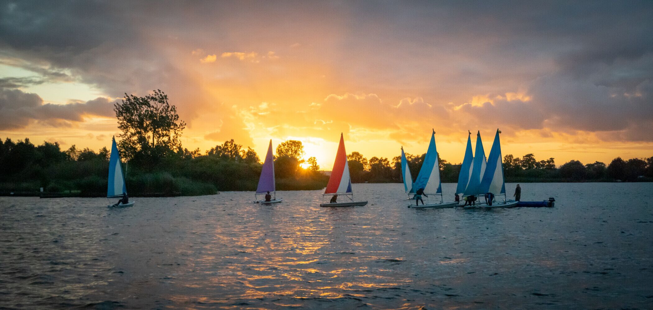 A sunset scene with multiple sailboats on calm water.