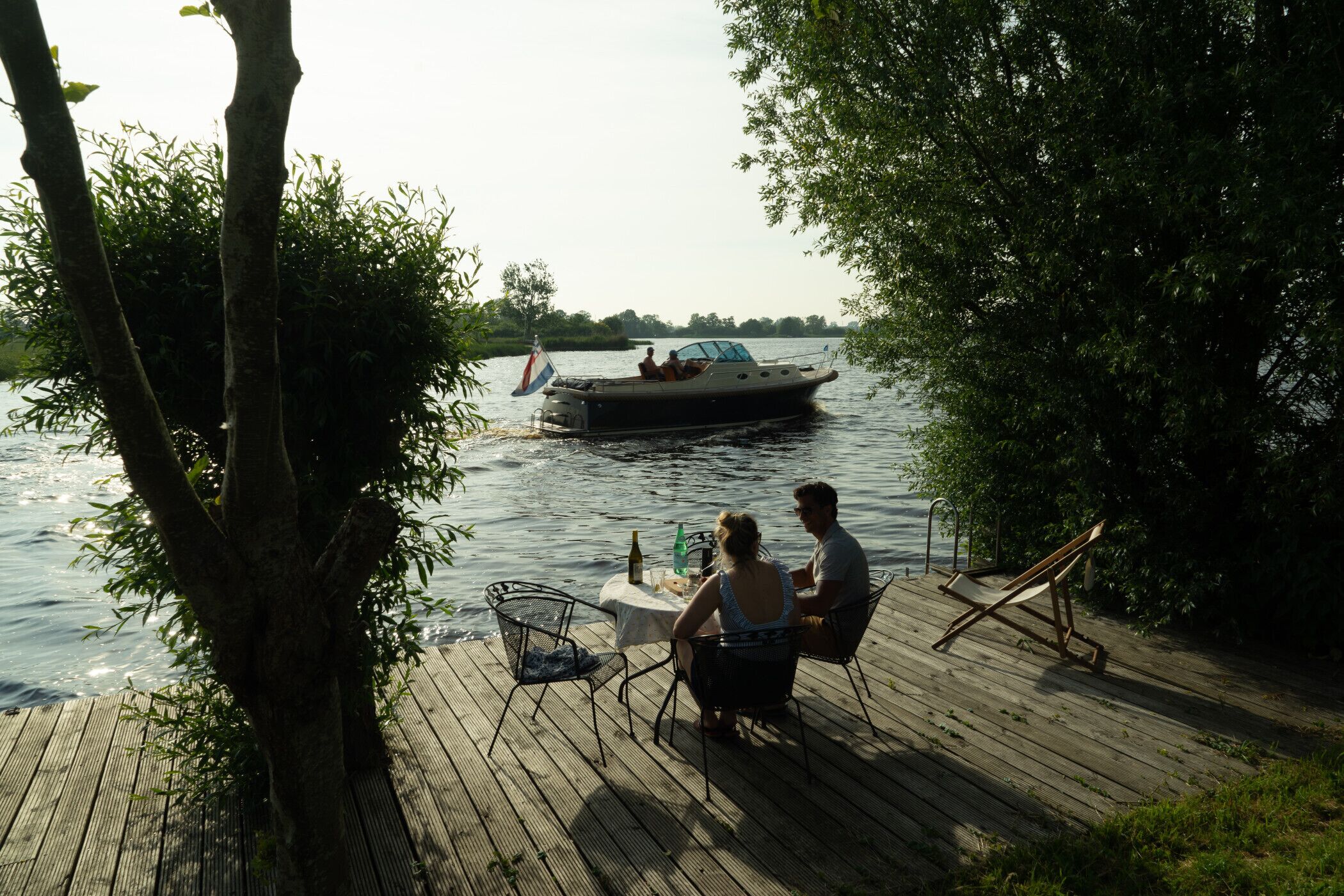 A wooden deck with a table and chairs, overlooking a calm river with a boat.