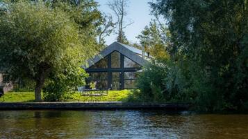A modern house with a large glass window, surrounded by lush greenery and a body of water.