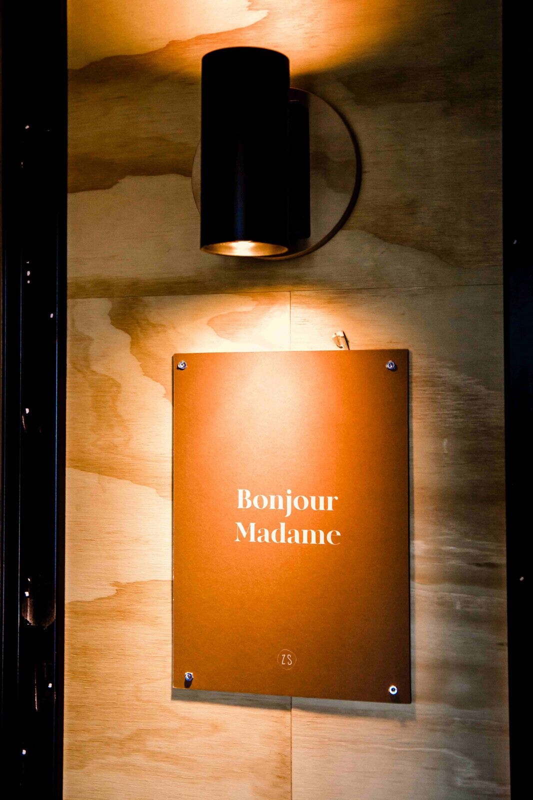 Une enseigne fixée au mur, portant l’inscription « Bonjour Madame » et un logo, et éclairée par un luminaire également fixé au mur.