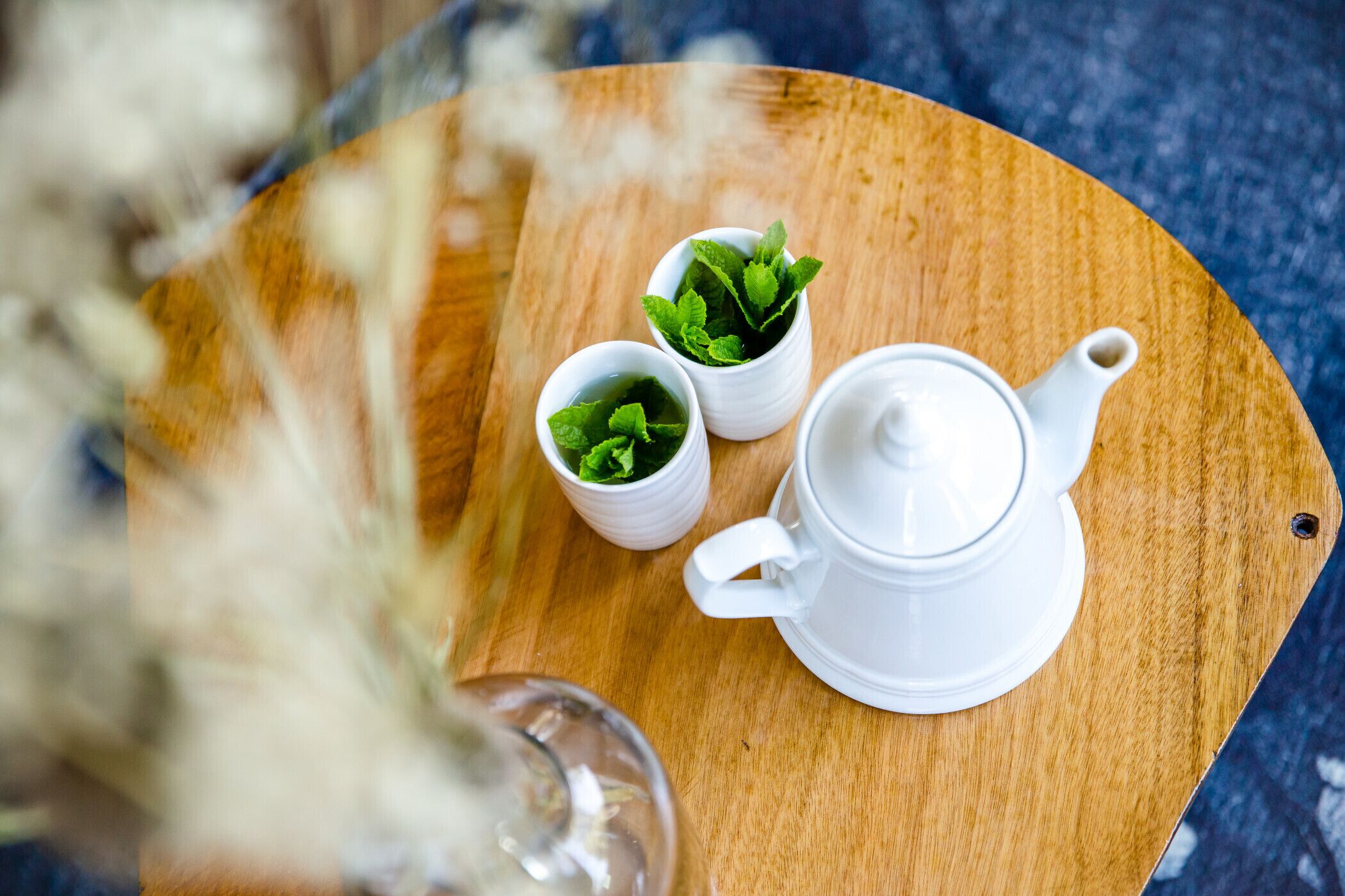 Une table en bois sur laquelle se trouvent une théière blanche et deux petites tasses blanches remplies de feuilles vertes, probablement de la menthe, posées sur une surface bleue texturée.