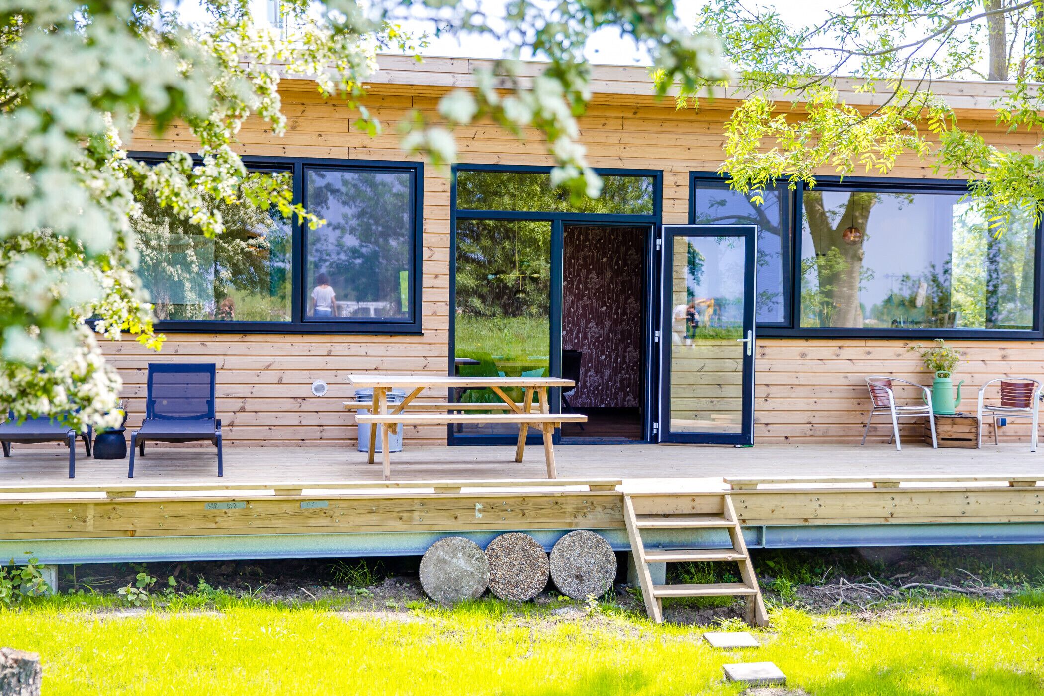 Une terrasse en bois avec une table de pique-nique et des chaises, de grandes fenêtres et une porte donnant sur un espace abrité.
