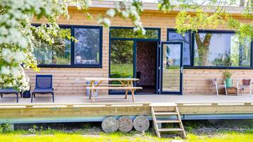 Une terrasse en bois avec une table de pique-nique et des chaises, de grandes fenêtres et une porte donnant sur un espace abrité.