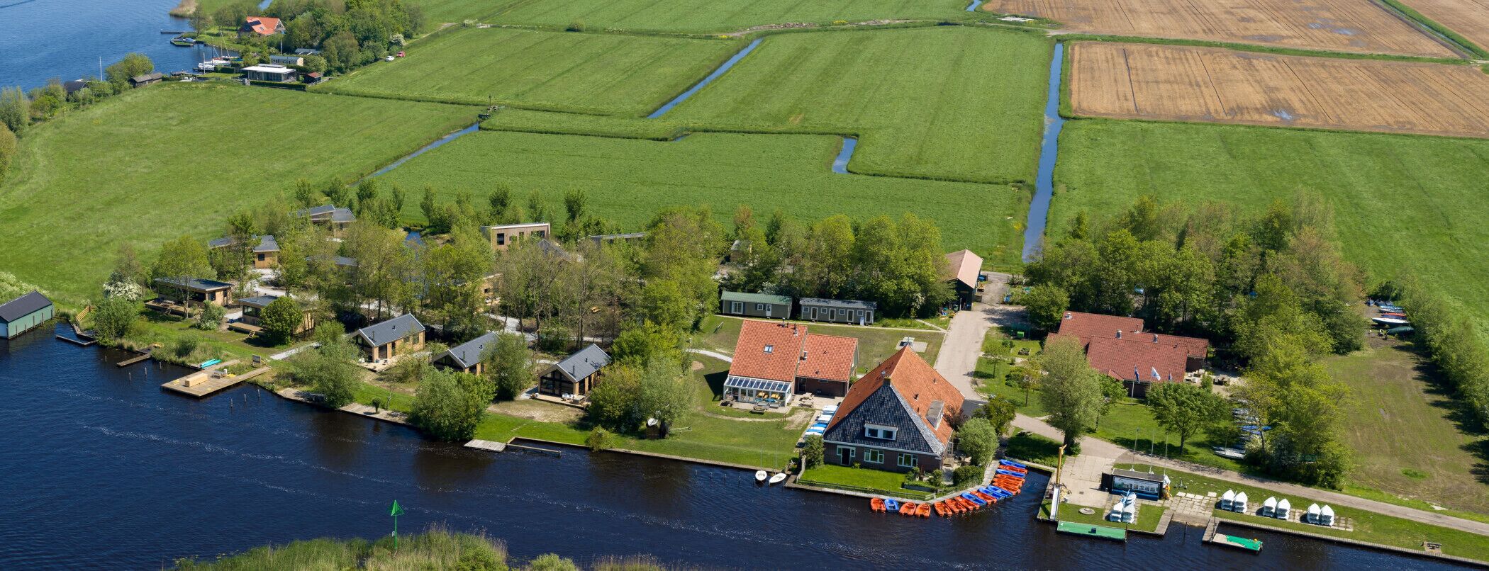 Luchtbeeld van een pand aan de rivier met huizen, een steiger met boten en groene velden.