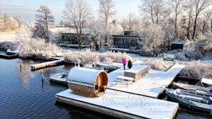 Escena invernal con un muelle que se adentra en un río helado, rodeado de árboles y edificios cubiertos de nieve.