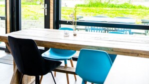 Comedor con mesa de madera y sillas azules, ventanas amplias con vista a follaje e iluminación con dos lámparas colgantes.