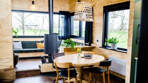 Une pièce chaleureuse avec un plancher en bois, une table ronde entourée de chaises, un canapé avec des coussins, une grande fenêtre avec des rideaux et un luminaire suspendu.