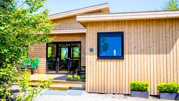 Un bâtiment moderne en bois, doté d’une grande entrée en verre, d’un espace extérieur aménagé pour s’asseoir et de plantes en pot.