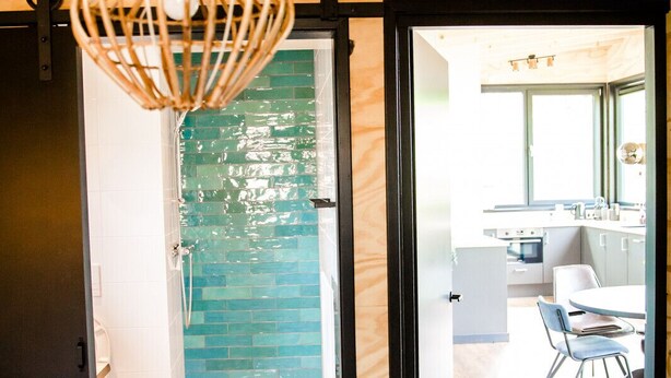 Un dormitorio con una cama, una lámpara colgante de mimbre, una puerta de vidrio verde que da al baño, y vistas a una cocina y comedor a través de una puerta abierta.
