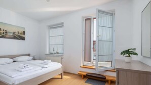 A modern bedroom with a wooden bed, a bench, a potted plant, and a view of the outdoors through a window. - Spacious Family Suite | Balcony | 2 Bedrooms (Swinoujscie)