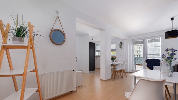 A modern living room with a wooden floor, white walls, a dining area with a white table and chairs, and a small kitchen visible in the background. - Spacious Family Suite | Balcony | 2 Bedrooms (Swinoujscie)