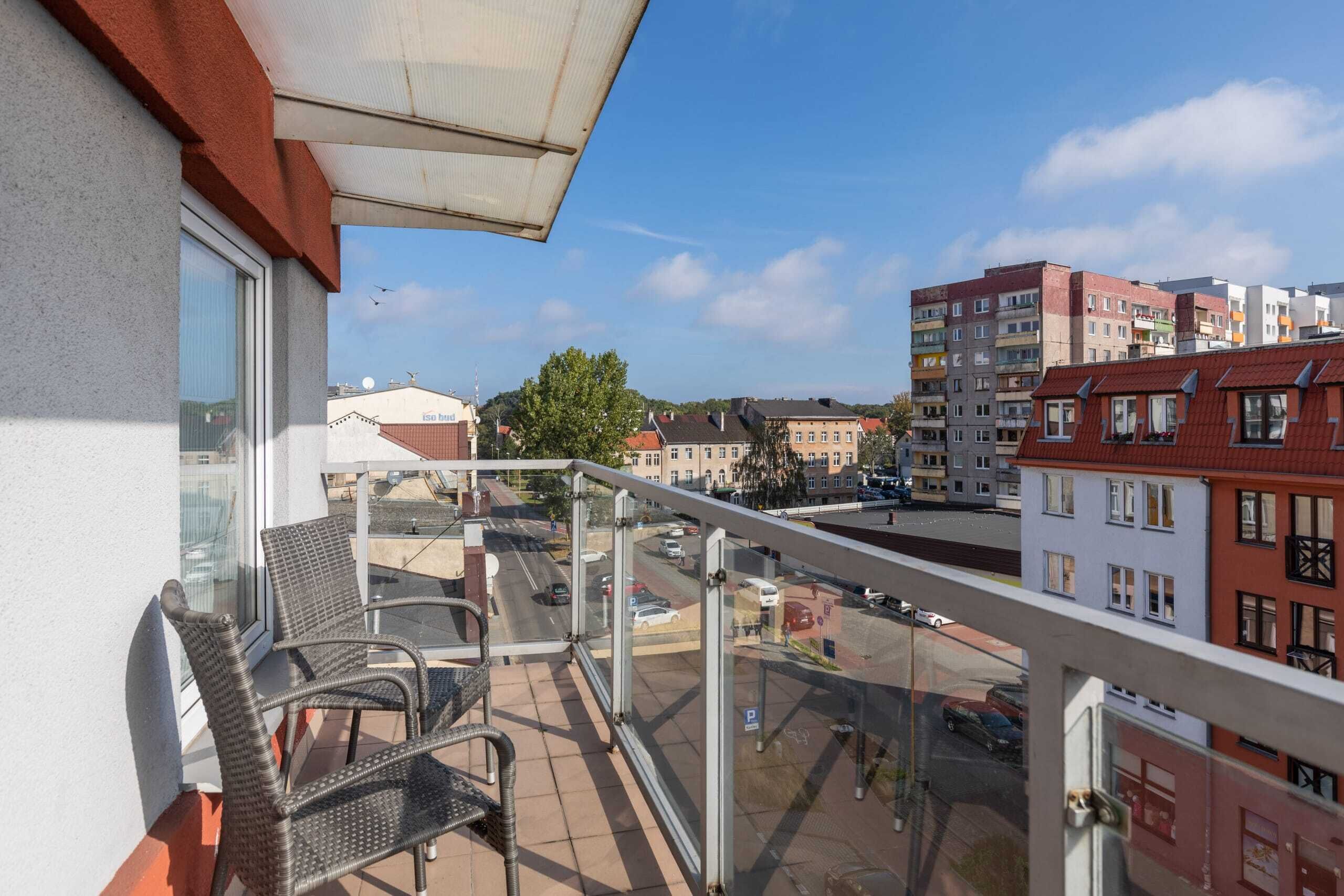 En balkong med flätade stolar, ett glasräcke och utsikt över en stadsgata med byggnader och parkerade bilar.