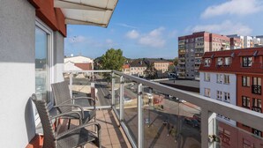 A balcony with wicker chairs, a glass railing, and a view of a city street with buildings and parked cars. - Spacious Family Suite | Balcony | 2 Bedrooms (Swinoujscie)