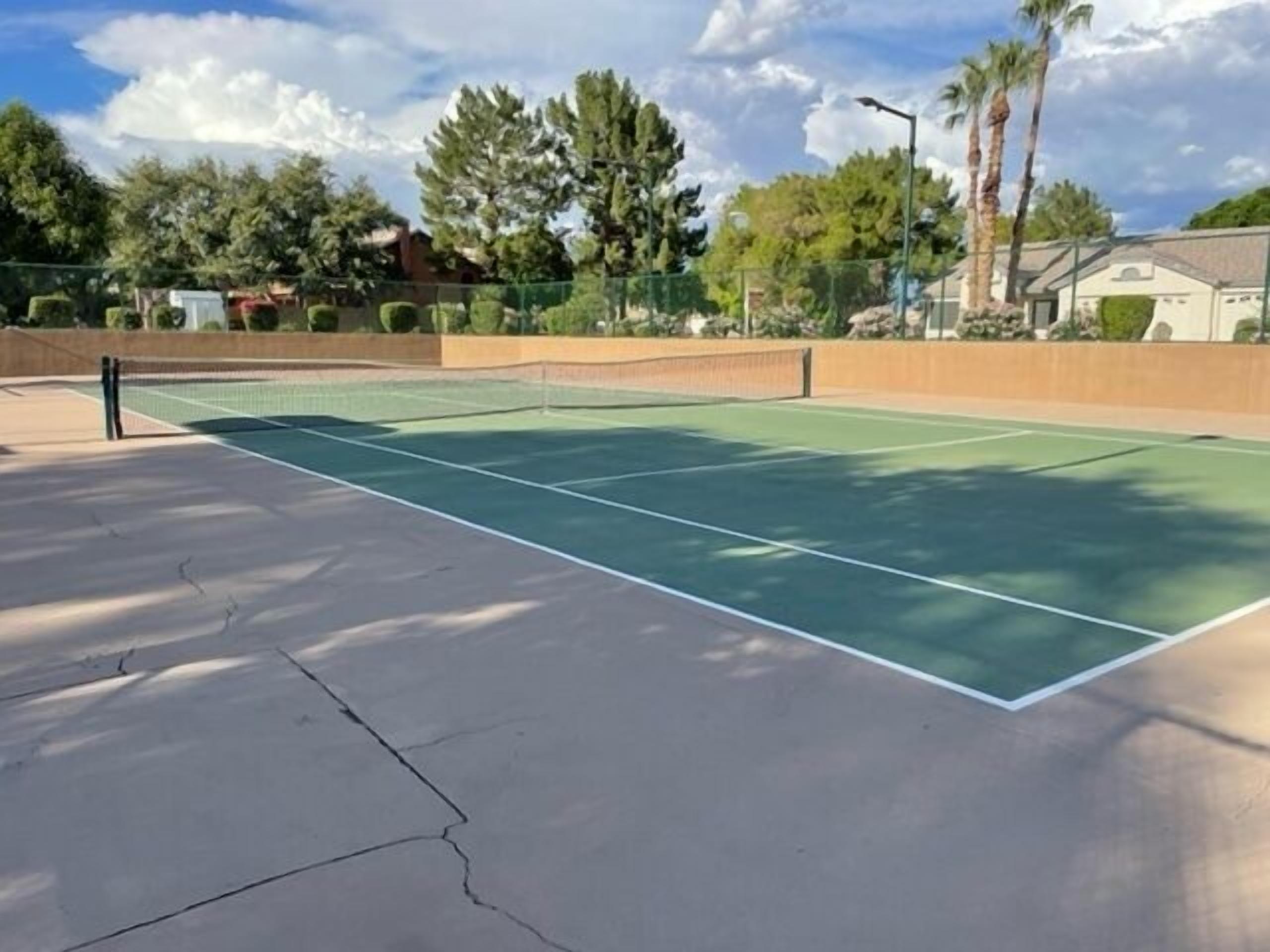 An outdoor tennis court with a net, surrounded by a fence, trees, and a building in the background.