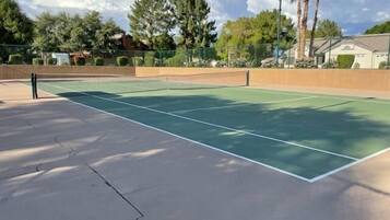 An outdoor tennis court with a net, surrounded by a fence, trees, and a building in the background.