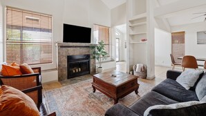 A modern living room with a fireplace, a flat-screen TV, and a large window with blinds. - Lakefront Home | Community Pool & Hot Tub (Chandler)