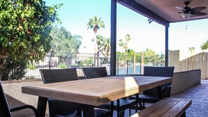 An outdoor patio with a wooden table and black wicker chairs, surrounded by a low fence and palm trees. - Lakefront Home | Community Pool & Hot Tub (Chandler)