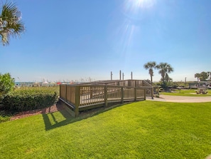 Una terraza de madera con barandilla, rodeada de césped cuidado y palmeras, con vistas a una playa.