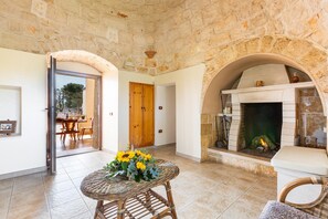 A room with stone walls, a fireplace, a dining area with a table and chairs, and an open door leading outside. - 2845 Trulli Carvaloo by Barbarhouse (Martina Franca)