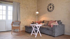 Una sala acogedora con un sofá gris, un sillón de mimbre, una mesita blanca y un reloj de pared.