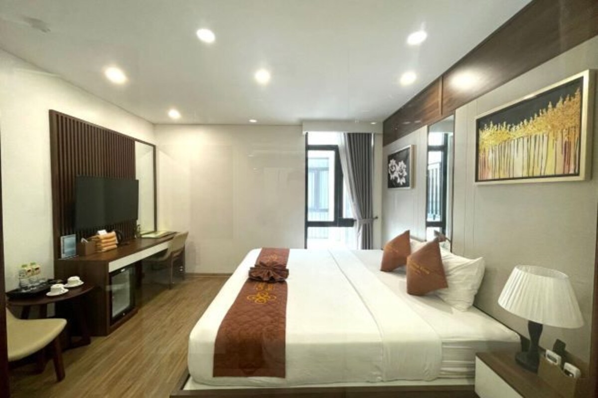 A hotel room with a bed, bedside table, lamp, TV, desk, and a window with curtains.