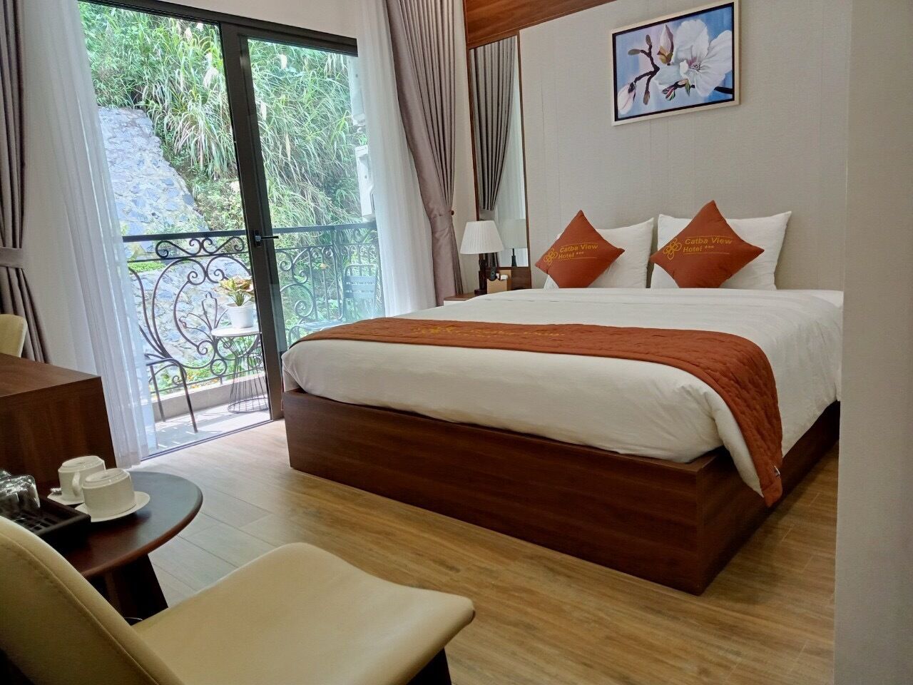A hotel room with a large bed, a balcony with a view of greenery, a desk with a chair, and a painting on the wall.