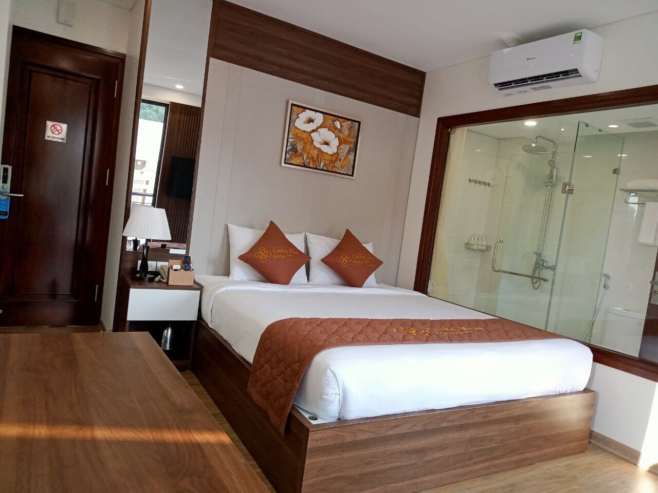 A hotel room with a wooden bed, a nightstand, a lamp, a painting on the wall, and a glass shower enclosure.