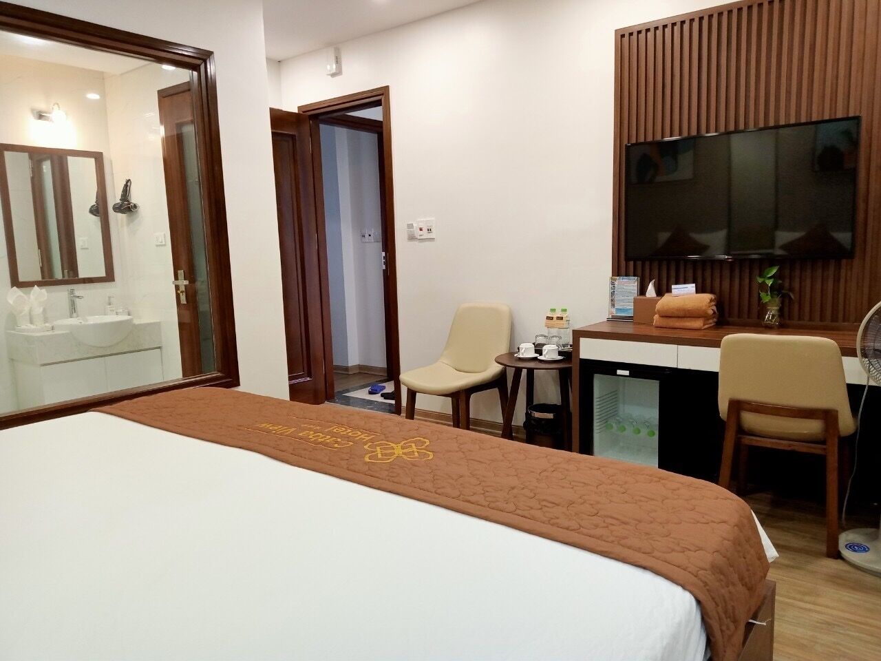Habitación de hotel con cama, escritorio, dos sillas, televisor y baño visible a través de una puerta abierta.