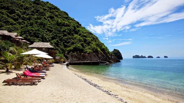 A beach with lounge chairs and umbrellas, a rocky cliff, and clear blue water. - Catba View Hotel (Hai Phong)