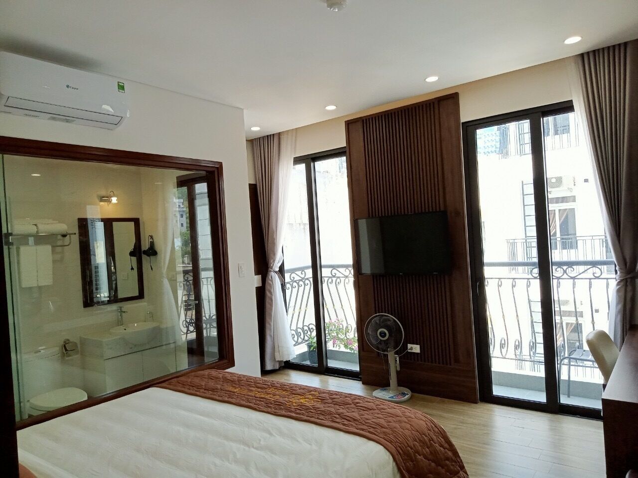 A hotel room with a large bed, a bathroom with a glass partition, a TV mounted on the wall, and a balcony with a view of the city.