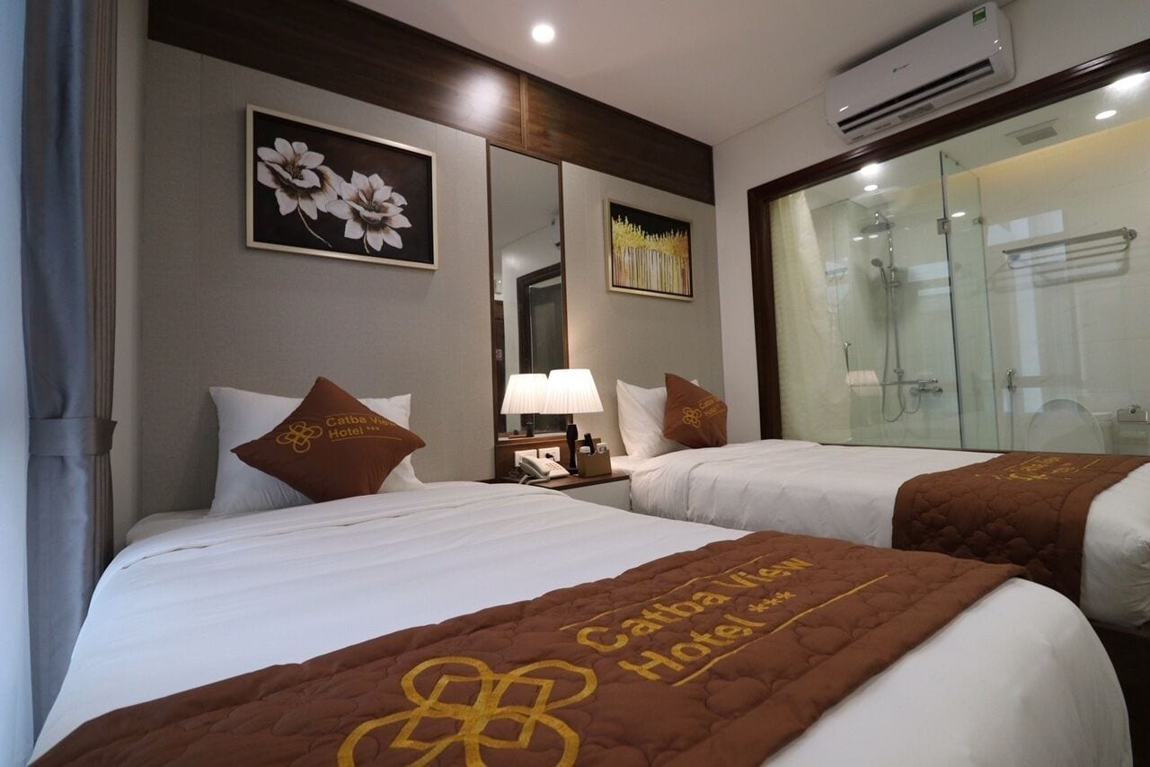 A hotel room with two beds, a nightstand with a lamp, a mirror, and a glass shower enclosure.