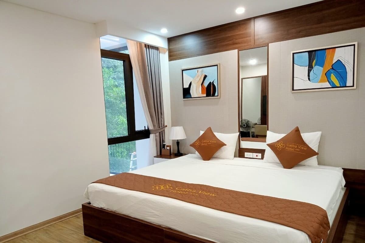 A hotel room with a large bed, a window with curtains, a mirror, and artwork on the walls.