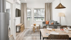 Une pièce moderne et bien éclairée, avec une table à manger dressée pour quatre personnes, un canapé, un téléviseur et une fenêtre donnant sur des arbres.
