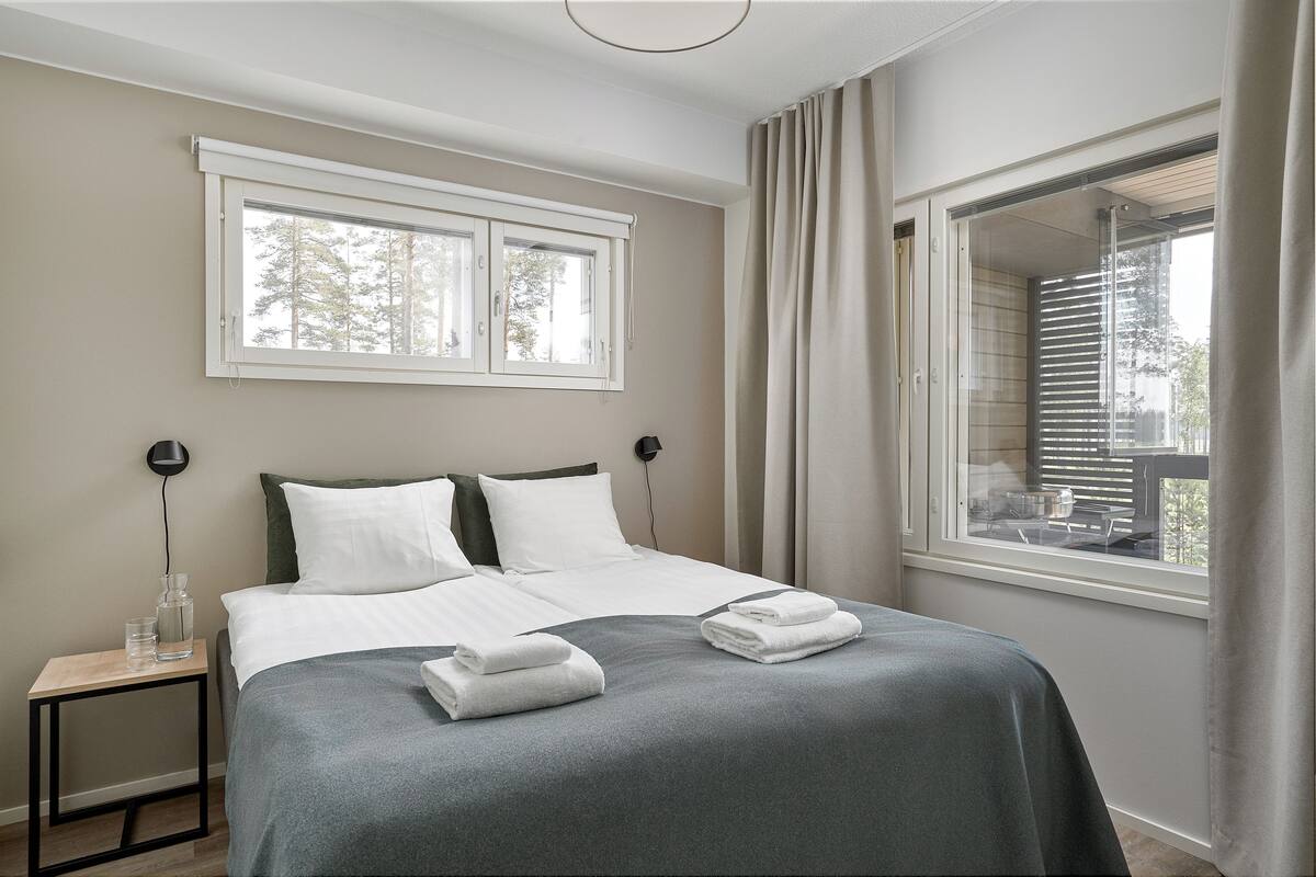 Een net opgemaakt bed met witte lakens, een nachtkastje met een lamp en een raam met blinds.