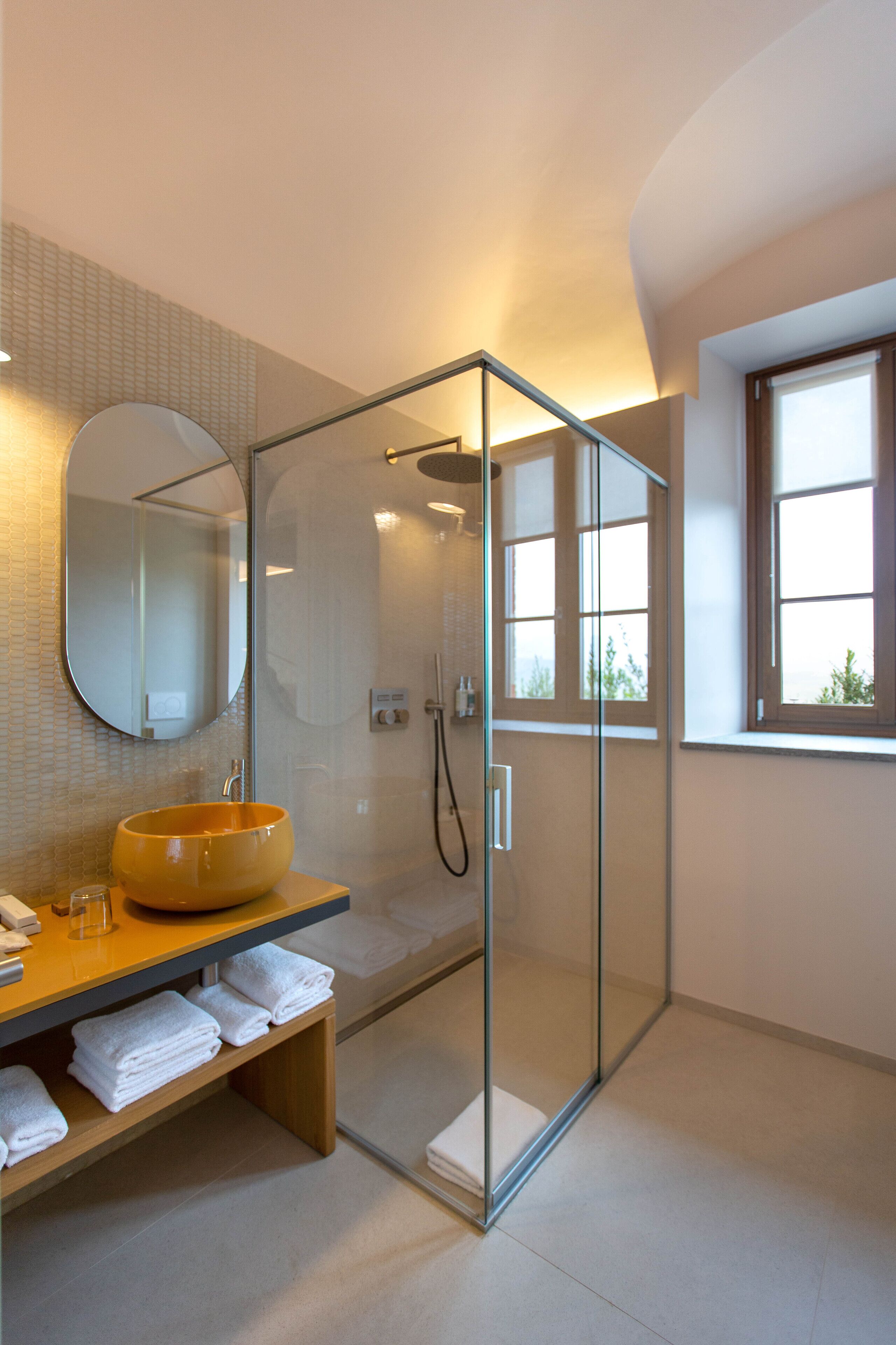 Een moderne badkamer met een douche met glazen wand, een rond wastafeltje en een grote spiegel.